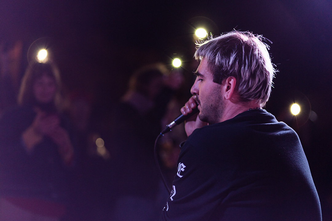 Concert photography of Salo. In the background a crowd is visible and some use the flashlight on their phones. The singer has a pensive look and holds the microphone with his right hand to his right cheek.