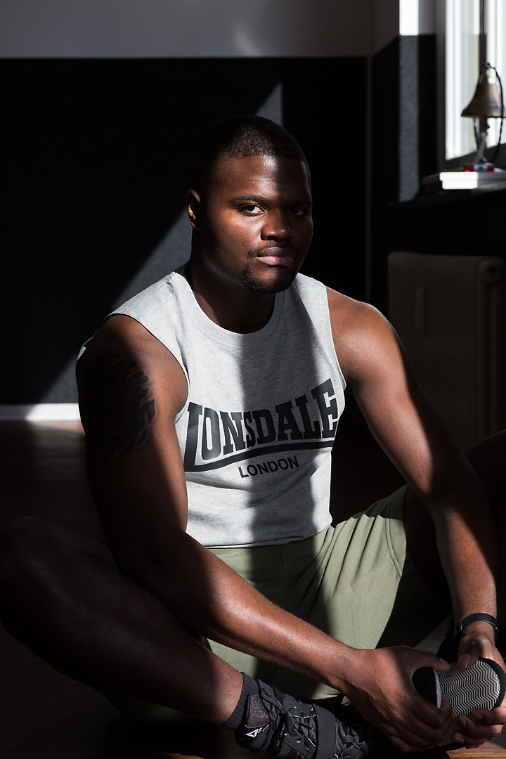 Portrait photography of Oluwaseun Salami, an Austrian professional boxer. He sits on a wooden gym floor and stretches. Half of his face is covered by the shadow of a window.