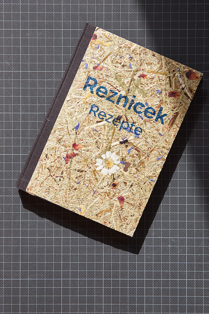 Photography of the cover design for a cook book. The hard cover is positioned on a black cutting board. The cover consists of pressed grass and flowers.